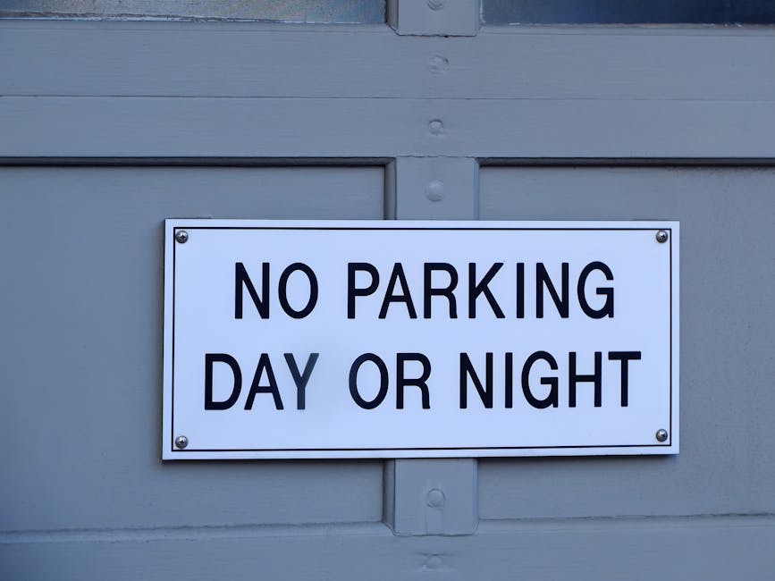 A rectangular white metal sign with bold black text reading 'NO PARKING DAY OR NIGHT' is mounted on a light grey wooden garage door with visible paneling and vertical grooves. The sign is secured with four screws, one at each corner. The environment appears to be during daylight hours, with natural lighting casting soft shadows on the door. The garage door shows signs of slight weathering, and the surroundings are clean and tidy. The image is relevant to house removals and moving logistics, as it depicts a typical parking restriction sign that may be encountered during a professional home relocation or furniture transport service provided by Man with Van Norwood Green, especially when adhering to local parking rules and skip regulations in Norwood Green.