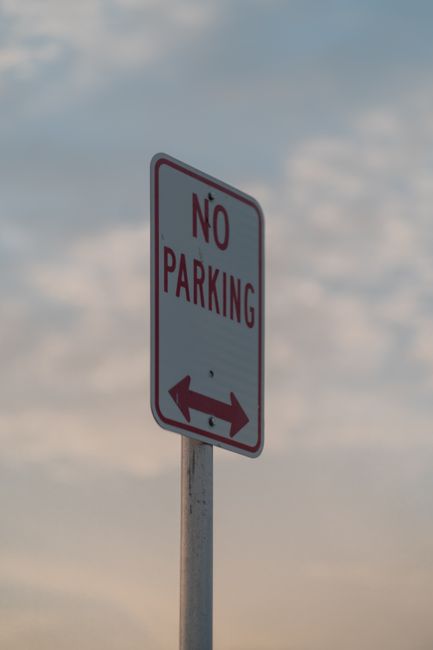 A tall, rectangular 'No Parking' sign with a white background, red border, and red text, mounted on a grey metal pole. The sign features a double-headed arrow at the bottom pointing left and right, indicating parking restrictions in both directions. The background shows a cloudy sky with patches of blue. This signage is situated near a property in Norwood Green, where house removals and furniture transport may be subject to parking and skip rules. The image emphasizes adherence to parking regulations, which are relevant to logistics and planning for home relocation services provided by Man with Van Norwood Green. The scene illustrates typical urban signage encountered during moving or packing and moving operations, supporting the need for careful planning when executing furniture transport and loading processes in residential areas.