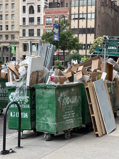 A row of three large green waste bins filled with assorted household rubbish and packaging materials is positioned outside a building on a city street, with some cardboard boxes, plastic wrapping, and metal fixtures overflowing from the bins. The area shows a commercial or residential urban environment with multi-storey buildings in the background, street lamps, and signage. A black metal pole with a retractable belt barrier is nearby, and a waste collection vehicle is visible in the distance. The presence of cardboard boxes, plastic sheeting, and bulky items indicates a home relocation or decluttering process as part of furniture transport or packing and moving activities. Man with Van Norwood Green provides professional removals and waste disposal services, supporting efficient house removals and logistics in the area.