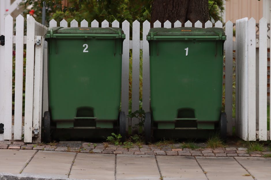 Two green wheeled rubbish bins numbered '1' and '2', positioned side by side in front of a white picket fence on a paved sidewalk, with a large tree trunk visible behind the fence. The bins are made of durable plastic, with lids closed, and are used for waste disposal as part of a home removal or moving logistics process. The surrounding area includes some small patches of grass and weeds growing between the paving stones, and the background features a garden with foliage. In the context of house removals and packing and moving services, these bins suggest waste collection or clearance activities associated with home relocation, handled by Man with Van Norwood Green during furniture transport and moving preparations.