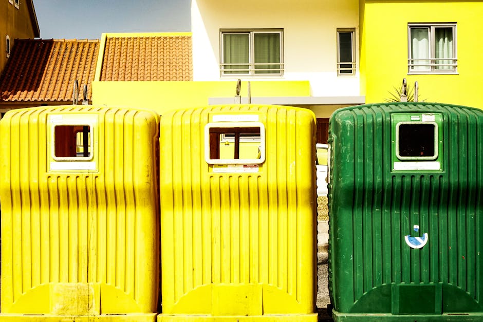 A row of three large green waste bins filled with assorted household rubbish and packaging materials is positioned outside a building on a city street, with some cardboard boxes, plastic wrapping, and metal fixtures overflowing from the bins. The area shows a commercial or residential urban environment with multi-storey buildings in the background, street lamps, and signage. A black metal pole with a retractable belt barrier is nearby, and a waste collection vehicle is visible in the distance. The presence of cardboard boxes, plastic sheeting, and bulky items indicates a home relocation or decluttering process as part of furniture transport or packing and moving activities. Man with Van Norwood Green provides professional removals and waste disposal services, supporting efficient house removals and logistics in the area.