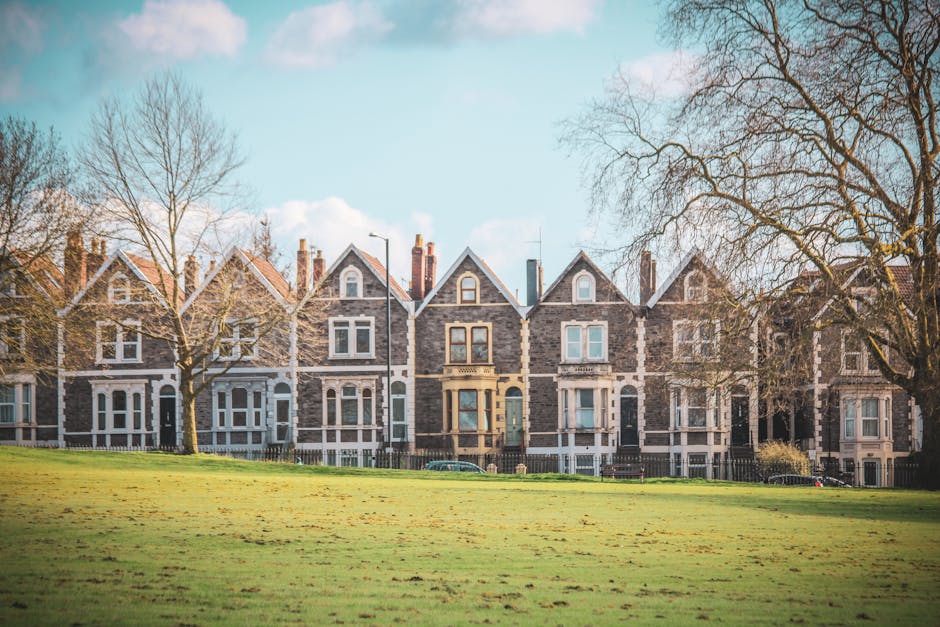 A row of Victorian-style terraced houses made of dark grey brick with white window frames and pitched roofs, situated behind a grassy park area with a few large, leafless trees. The houses feature bay windows on the ground and first floors, small front gardens separated by low fences, and some with chimneys on the rooftops. The image is taken on a clear day with a blue sky and a few scattered clouds, providing natural lighting. In front of the houses, there are paving stones and a few parked cars visible along the street. This scene depicts an urban residential neighbourhood, typical of a home relocation or furniture transport context, with a focus on the front facades of the houses and the surrounding environment, as seen from the park area.