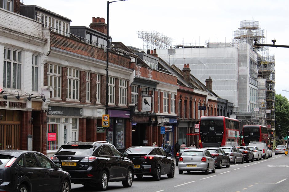 A busy street scene in Norwood Green with a row of traditional brick terraced houses, some under scaffolding indicating ongoing building work, and parked cars lining the roadside. Two red double-decker buses are visible, with a black SUV and several other vehicles parked along the curb. Pedestrians are walking on the pavement, and the environment appears to be a residential and commercial area with shops and businesses on the ground floors of the buildings. The scene is captured during daylight with overcast weather, and evidence of moving or loading activity is not directly visible in this image. Man with Van Norwood Green provides house removal and furniture transport services, and this street showcases a typical setting where such services may be required for home relocation or moving logistics.