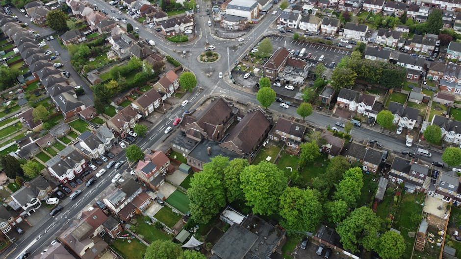 A quiet residential street with a narrow paved roadway leading to a row of traditional brick terraced houses featuring chimney stacks and pitched roofs. On the left side, a low brick wall and a wooden
