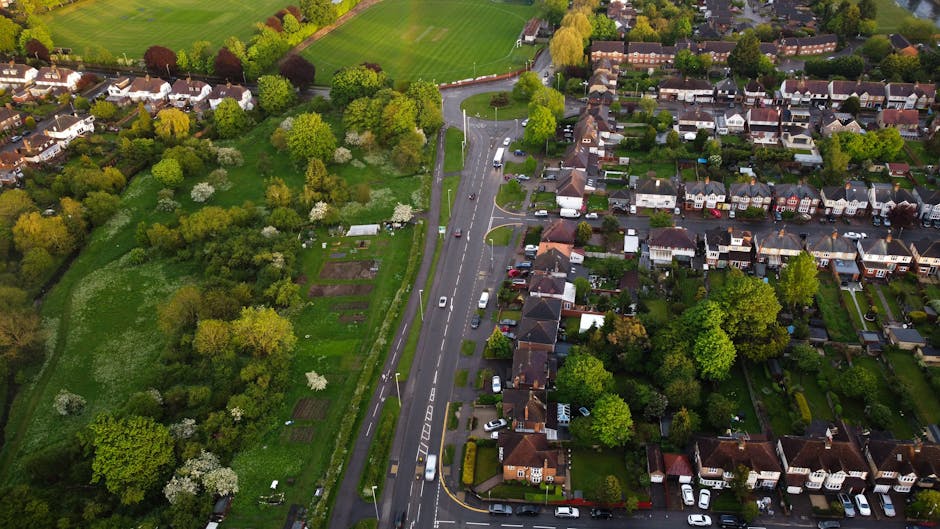 An aerial view of a residential neighbourhood in Norwood Green showing a main road lined with parked cars and a variety of houses with front gardens, surrounded by lush green trees and open green spaces. The image captures the calm, suburban atmosphere during daylight with natural lighting highlighting the foliage and residential buildings. This scene exemplifies a typical home environment where house removals and furniture transport may take place, with close proximity of properties and local roads suitable for relocation logistics managed by companies like Man with Van Norwood Green.