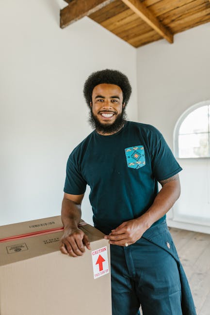 A man with dark skin and an afro hairstyle stands inside a bright, well-lit room with white walls and a wooden ceiling. He is smiling and wearing a dark blue T-shirt with a small patterned pocket on the left side. The man is holding a large cardboard box with both hands, which has a red and white 'CAUTION' arrow label on the side. The room features a rounded window in the background, letting in natural light, and appears to be part of a home or property preparing for moving or furniture transport. This scene reflects home relocation activities, with the man associated with professional removals services such as those offered by Man with Van Norwood Green, supporting residential moving and packing processes.