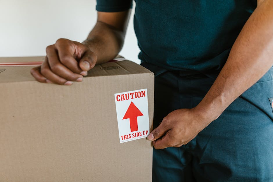 Close-up of a worker's hands securing a cardboard box during a home relocation process, with the worker wearing a dark blue uniform. The box is sealed with packing tape and features a red and white caution label that reads 'CAUTION THIS SIDE UP' with an upward-pointing arrow, indicating proper orientation for handling. The worker is inside a property, possibly near a doorway or in a room prepared for packing and moving, and is actively involved in the furniture transport and packing process. In the background, minimal details are visible, focusing on the box and the worker's hands. This scene reflects the careful handling and logistics involved in professional removals, such as those provided by Man with Van Norwood Green, supporting efficient house removals and moving services.