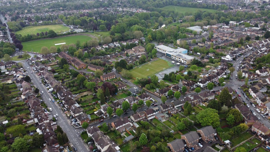An aerial view of a residential neighbourhood in Norwood Green showing rows of houses with garden lawns and trees along the streets. In the center of the image, a sports field with green grass is visible, surrounded by a few sports buildings. Nearby, a modern, white commercial or school building with large windows is situated. The streets are lined with parked cars, and some are actively being moved or loaded onto a van. The environment is lush with mature trees and greenery, with surrounding open fields and further housing visible in the distance under an overcast sky. This scene captures the setting for a home relocation or furniture transport process, reflecting typical moving logistics handled by Man with Van Norwood Green, as part of their comprehensive removals services linked to house moves in the area.