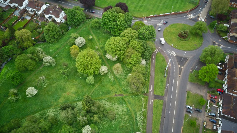 An aerial view of a residential neighbourhood in Norwood Green showing a large green park with various mature trees, some with white blossoms, bordered by a narrow footpath. To the right, a roundabout connects multiple roads, with vehicles including cars and vans visible on the junction and adjacent streets. On the far side of the park, rows of terraced houses with pitched roofs, brick facades, and front gardens are visible. The scene is lit by natural daylight, highlighting the lush greenery and the streets' traffic flow, illustrating a typical home relocation and moving logistics environment supported by [COMPANY_NAME] for efficient furniture transport and packing during house removals.