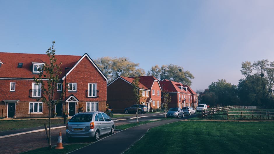 An aerial view of a residential neighbourhood in Norwood Green showing rows of houses with garden lawns and trees along the streets. In the center of the image, a sports field with green grass is visible, surrounded by a few sports buildings. Nearby, a modern, white commercial or school building with large windows is situated. The streets are lined with parked cars, and some are actively being moved or loaded onto a van. The environment is lush with mature trees and greenery, with surrounding open fields and further housing visible in the distance under an overcast sky. This scene captures the setting for a home relocation or furniture transport process, reflecting typical moving logistics handled by Man with Van Norwood Green, as part of their comprehensive removals services linked to house moves in the area.