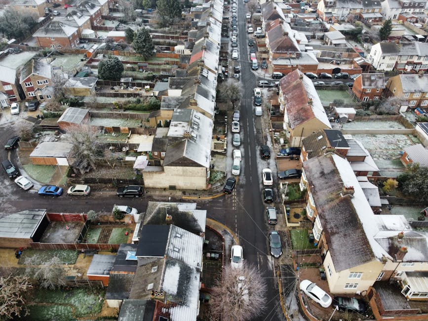 An aerial view of a residential street in Norwood Green during winter, showing terraced houses with varied roof styles, some with snow on the rooftops and garden sheds, and cars parked along the narrow, wet road. The street is lined with trees and fences, and frost covers grass lawns and driveways. In the foreground, a house with a small backyard features a shed and garden furniture, while the road extends into the distance with additional parked vehicles. This image illustrates a typical suburban neighbourhood where home relocation and furniture transport services by Man with Van Norwood Green may be required for a smooth move day and effective packing and moving logistics.