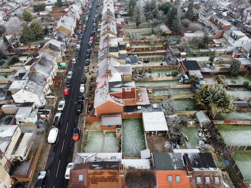 An aerial view of a residential street in Norwood Green during winter, showing terraced houses and detached homes with snow-covered roofs. The street is lined with parked cars, including sedans, hatchbacks, and vans. Several back gardens are visible, enclosed by wooden fences, some with sheds, garden furniture, and trimmed trees. The houses have different roofing materials such as tiles and felt, with some rooftops covered in snow. In the foreground, a terraced house has a driveway next to it and a small yard with a shed, while other properties feature lawns or paved areas. The environment is quiet, with no visible people or ongoing activity, depicting a typical suburban neighborhood setting. This image illustrates the typical conditions for home relocation and furniture transport in Norwood Green, with an emphasis on residential property layouts and outdoor spaces suitable for removals services by Man with Van Norwood Green.