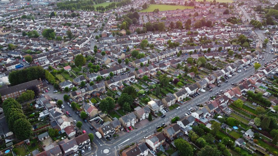 An aerial view of a residential street in Norwood Green during winter, showing terraced houses with varied roof styles, some with snow on the rooftops and garden sheds, and cars parked along the narrow, wet road. The street is lined with trees and fences, and frost covers grass lawns and driveways. In the foreground, a house with a small backyard features a shed and garden furniture, while the road extends into the distance with additional parked vehicles. This image illustrates a typical suburban neighbourhood where home relocation and furniture transport services by Man with Van Norwood Green may be required for a smooth move day and effective packing and moving logistics.