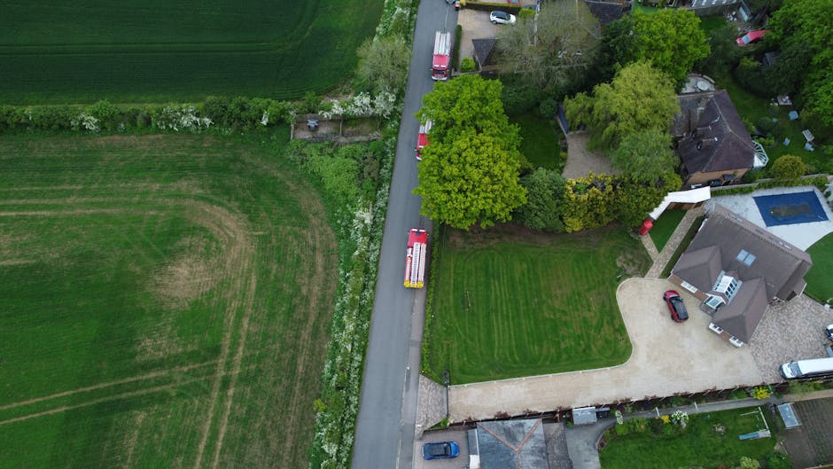 An aerial view of a suburban residential property showing a house with a paved driveway and a neatly maintained lawn, surrounded by trees and gardens. A red van with a white cab, used for home relocation and furniture transport, is parked along the narrow grey asphalt street adjacent to the property, partially obstructing the entrance. The van is positioned close to the front yard, indicating the loading process for packing and moving activities. Nearby, a red car is parked in the driveway near the house, with another vehicle visible closer to the street. The surrounding area includes other houses with similar features such as patios and swimming pools, situated amidst verdant gardens and trees. The scene captures the typical environment for same-day removals in Norwood Green, with clear indications of ongoing furniture transport and packing preparations within a well-maintained neighbourhood.