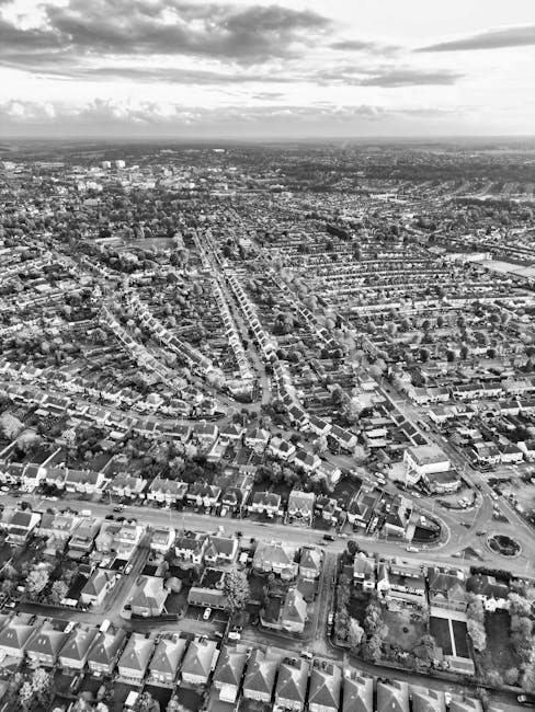 A black-and-white aerial photograph of a densely populated residential area in Norwood Green, showing numerous single-family houses with pitched roofs, arranged along curving streets and cul-de-sacs. In the foreground, there are several detached and semi-detached homes with front gardens, driveways, and parked cars. The streets are lined with trees and some open green spaces are visible amidst the housing. The background extends towards the horizon with an expansive view of the town and distant horizon under partly cloudy skies. This image captures the typical layout of a suburban neighbourhood, relevant to home relocation and furniture transport services provided by Man with Van Norwood Green during house removals, especially in the context of packing, logistics, and loading at the property or onto a van for transportation.