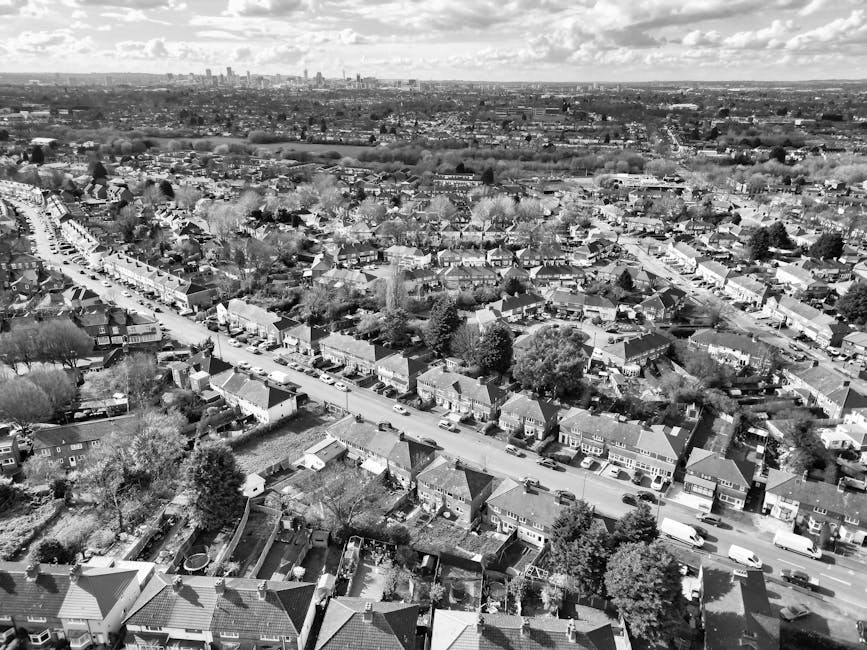 An aerial view of a suburban residential property showing a house with a paved driveway and a neatly maintained lawn, surrounded by trees and gardens. A red van with a white cab, used for home relocation and furniture transport, is parked along the narrow grey asphalt street adjacent to the property, partially obstructing the entrance. The van is positioned close to the front yard, indicating the loading process for packing and moving activities. Nearby, a red car is parked in the driveway near the house, with another vehicle visible closer to the street. The surrounding area includes other houses with similar features such as patios and swimming pools, situated amidst verdant gardens and trees. The scene captures the typical environment for same-day removals in Norwood Green, with clear indications of ongoing furniture transport and packing preparations within a well-maintained neighbourhood.