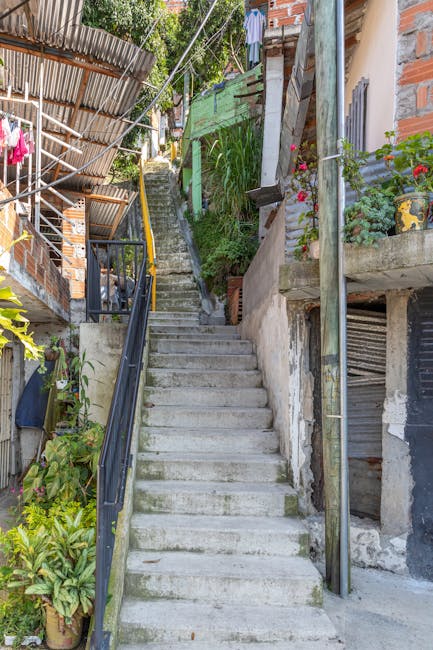 A concrete outdoor staircase with multiple steps ascends through a narrow, residential area in Norwood Green, featuring a black metal handrail on the left side. To the right, there is a concrete support pillar and exposed brickwork, along with various potted plants and flowers on small concrete and wooden ledges attached to the building exterior. Some steps show slight staining and wear, with greenery and leafy plants growing along the sides and at the top of the stairs. A green painted fence and wooden panels are visible at the upper part of the stairs, along with trees and additional foliage in the background, providing natural sunlight and shadows on the scene. This setup suggests a home relocation or furniture transport process in a densely built residential area, with [COMPANY_NAME] likely involved in moving services through narrow streets and staircase access points.
