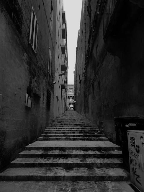 A narrow outdoor staircase with concrete steps leading upward between tall, closely positioned brick buildings in Norwood Green. The steps show signs of wear and some graffiti is visible on a utility box at the base. The scene is captured in black and white, with diffuse, overcast lighting suggesting cloudy weather. This setting reflects an urban environment requiring careful navigation during home relocation or furniture transport. The image highlights the challenging access aspect that Man with Van Norwood Green’s removals service can address when planning moves involving narrow streets and staircases in residential areas, emphasizing the importance of professional packing and moving logistics in constrained spaces.